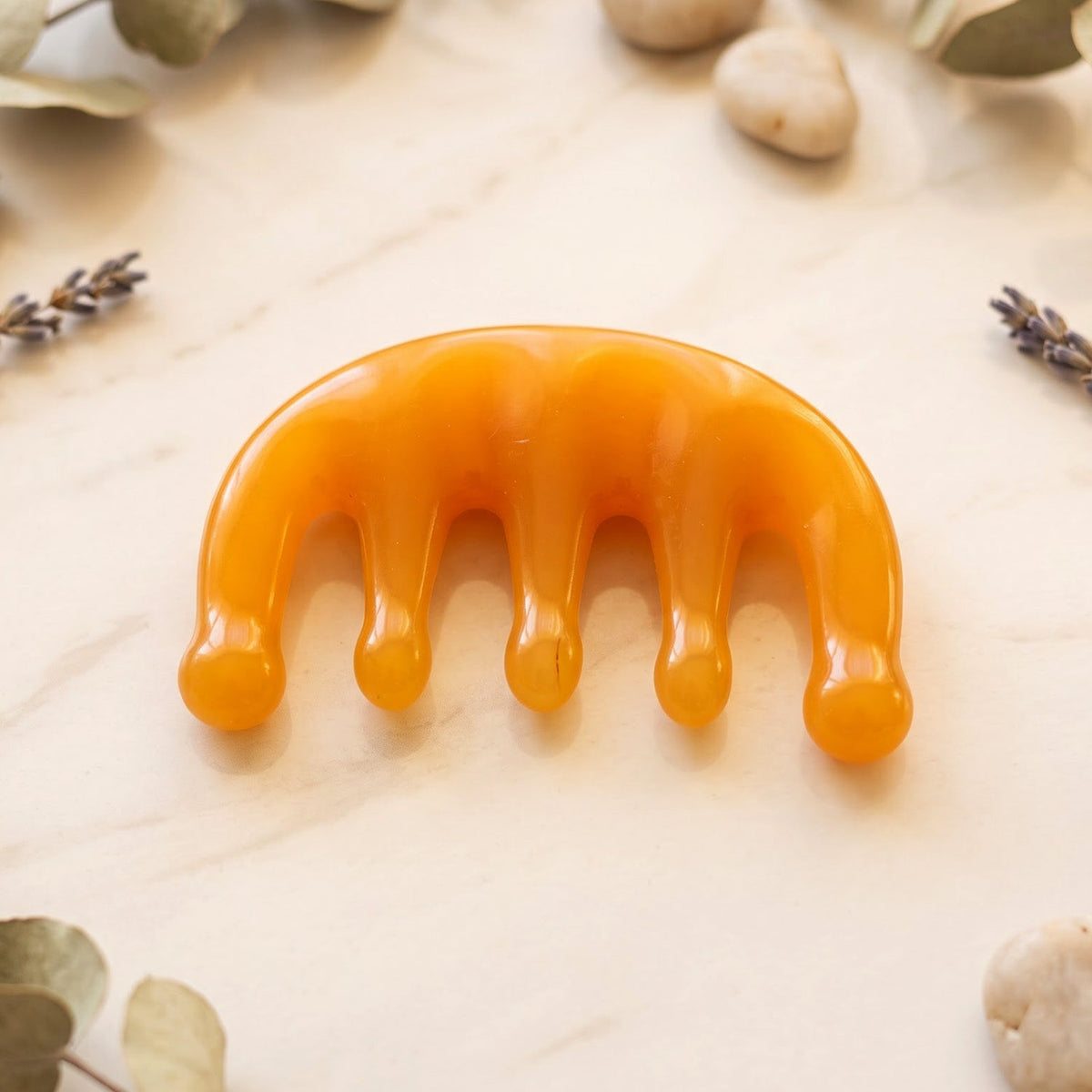The orange scalp massager comb is centered on a white marble surface, surrounded by dried eucalyptus leaves and lavender