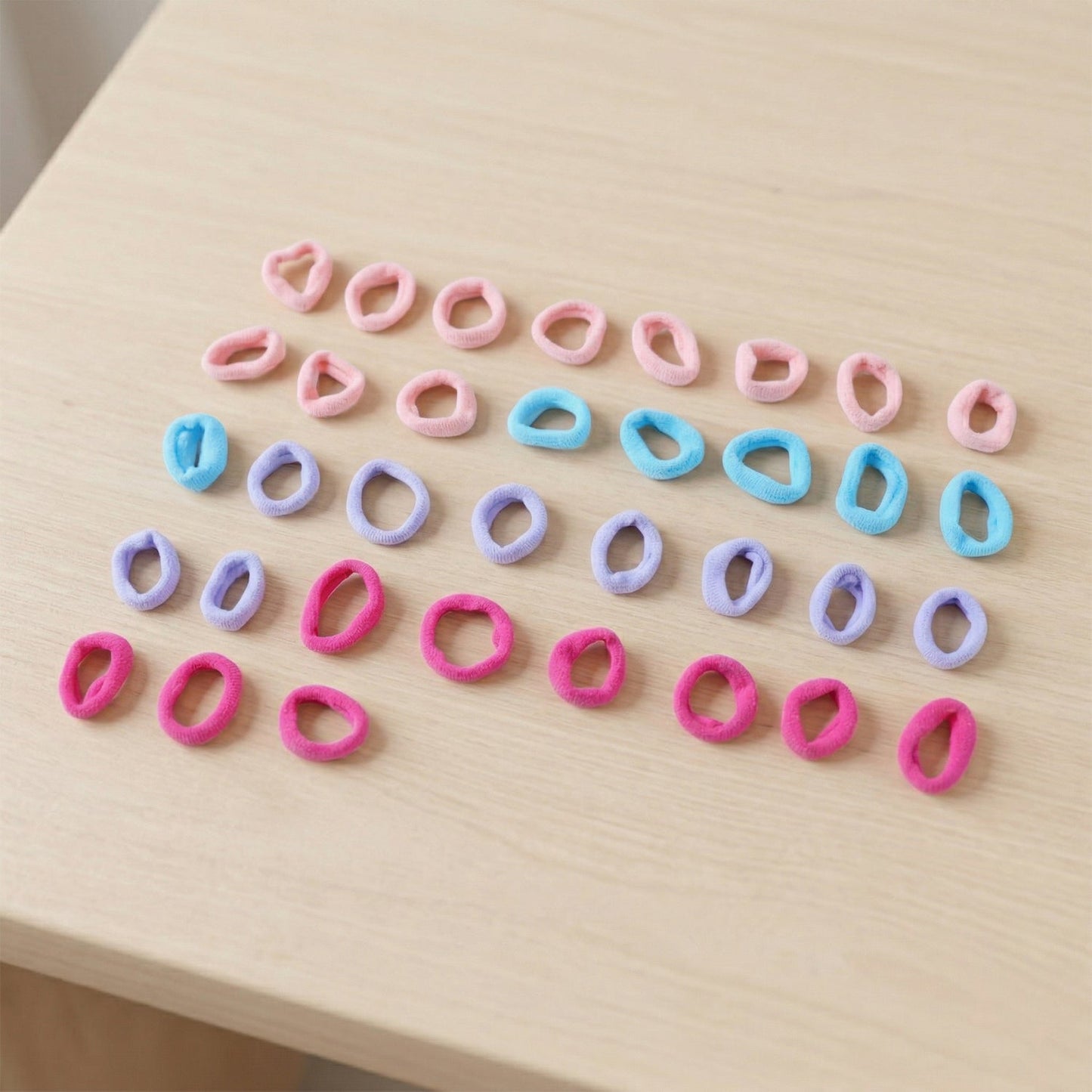 Thirty-five assorted pink, blue, lavender, and fuchsia elastic hair ties scattered on a light wooden surface.