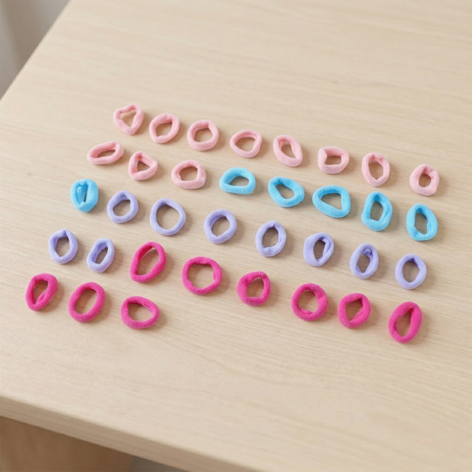 Thirty-five assorted pink, blue, lavender, and fuchsia elastic hair ties scattered on a light wooden surface.