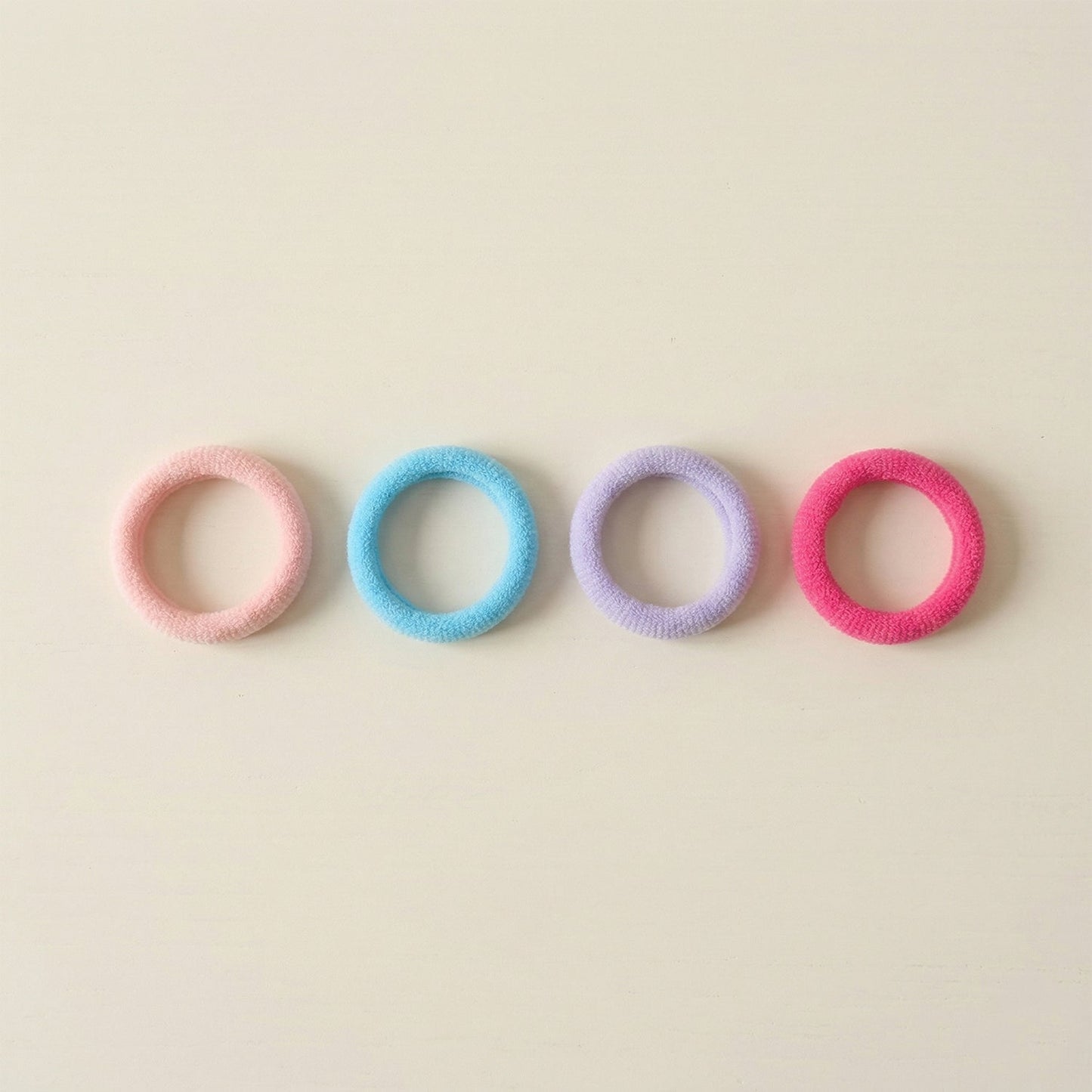 Four colorful elastic hair ties in pink, blue, lavender, and fuchsia arranged in a row on a light beige surface.