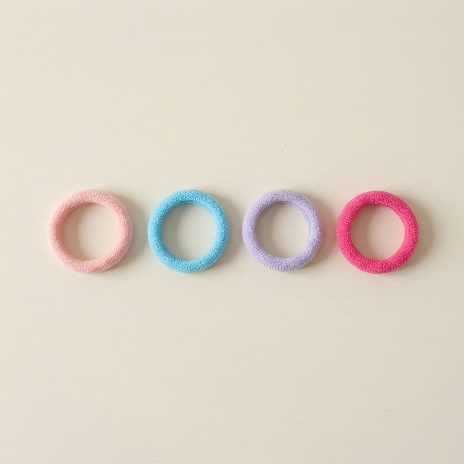 Four colorful elastic hair ties in pink, blue, lavender, and fuchsia arranged in a row on a light beige surface.