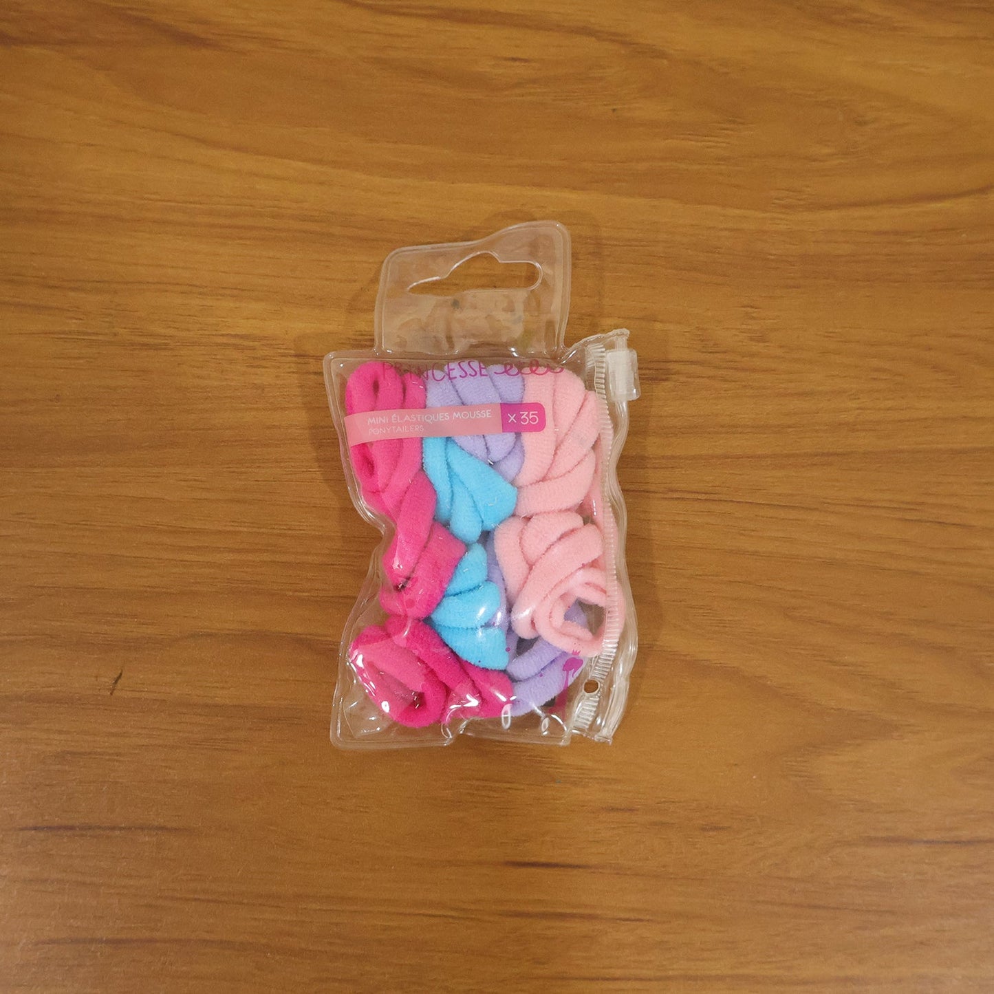 A clear plastic package containing 35 colorful elastic hair ties, including pink, blue, and lavender, on a wooden surface.