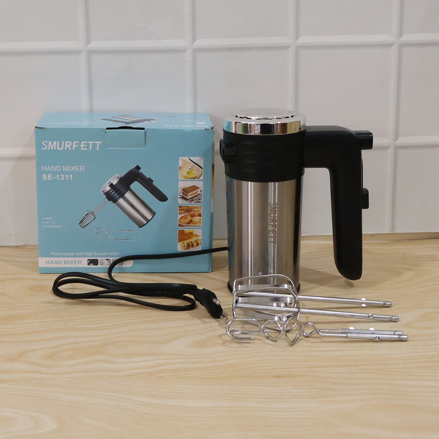 A light blue product box and a black and silver electric hand mixer with beaters and dough hooks are on a light wooden