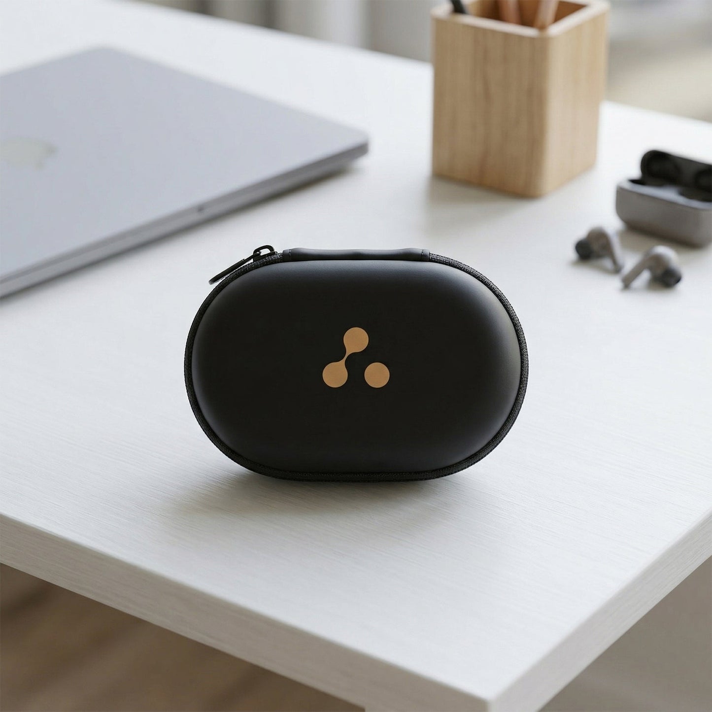 A closed black hard shell case with a gold three-dot logo sits on a white desk next to a laptop and pencil holder.