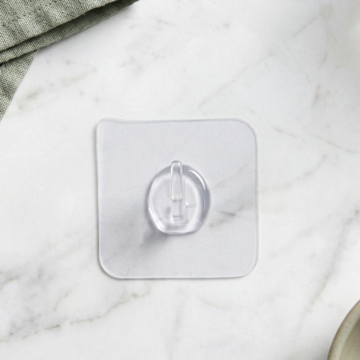 A clear self-adhesive wall hook centered on a light grey marble surface with a green cloth in the corner.
