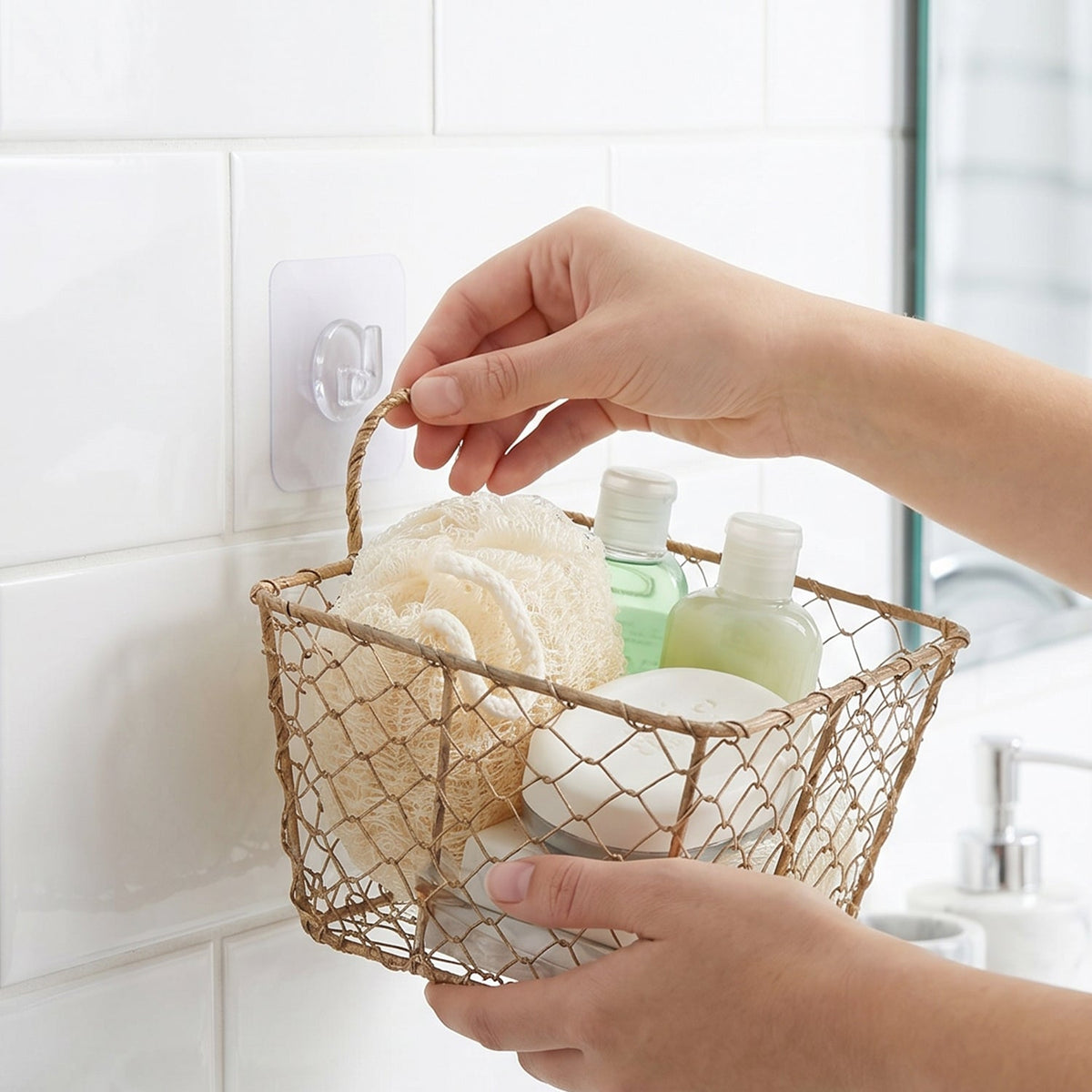 Hands holding a wire basket filled with bath products, hanging from a clear wall hook on white tiles.