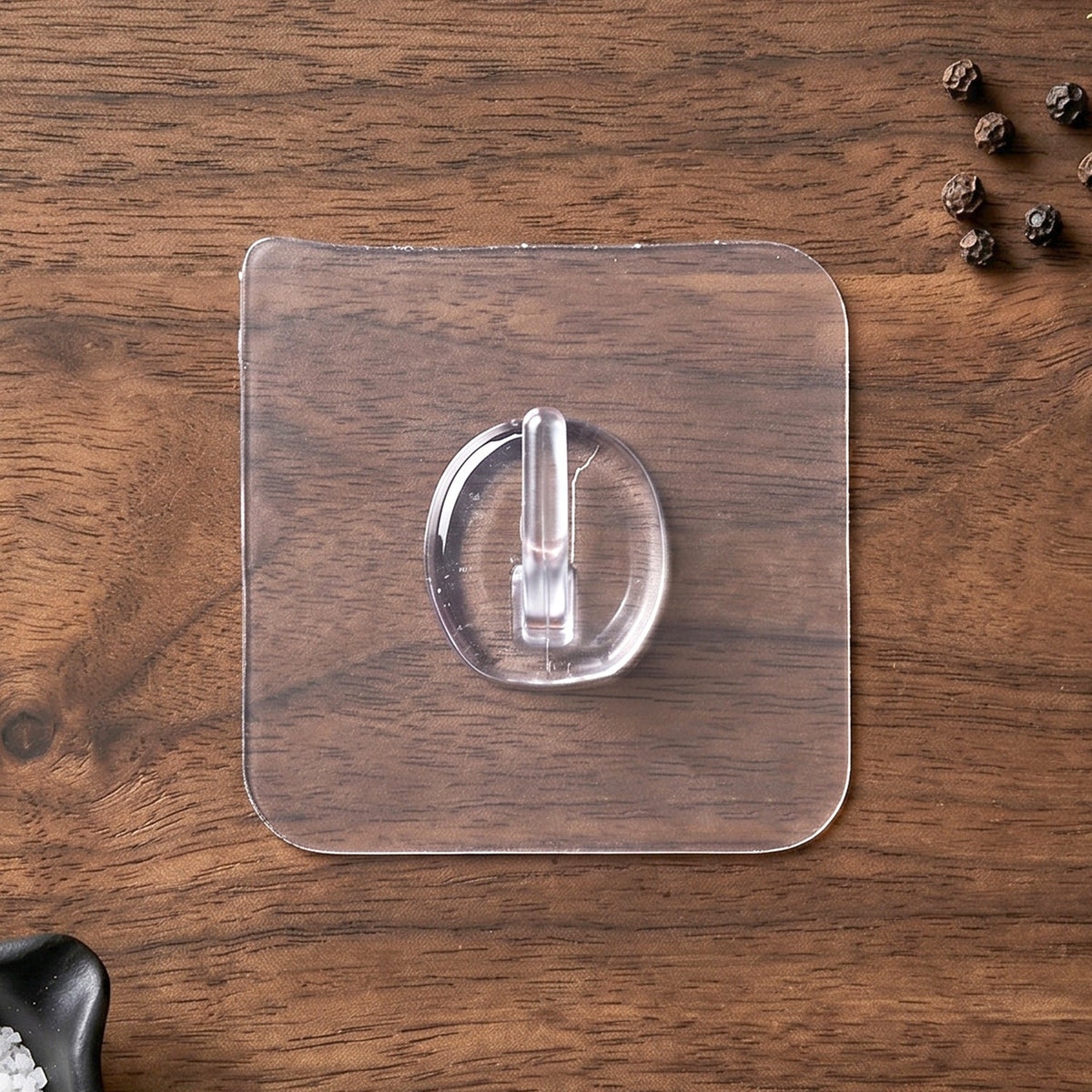 A clear self-adhesive wall hook on a dark wooden surface, with black peppercorns scattered in the background.