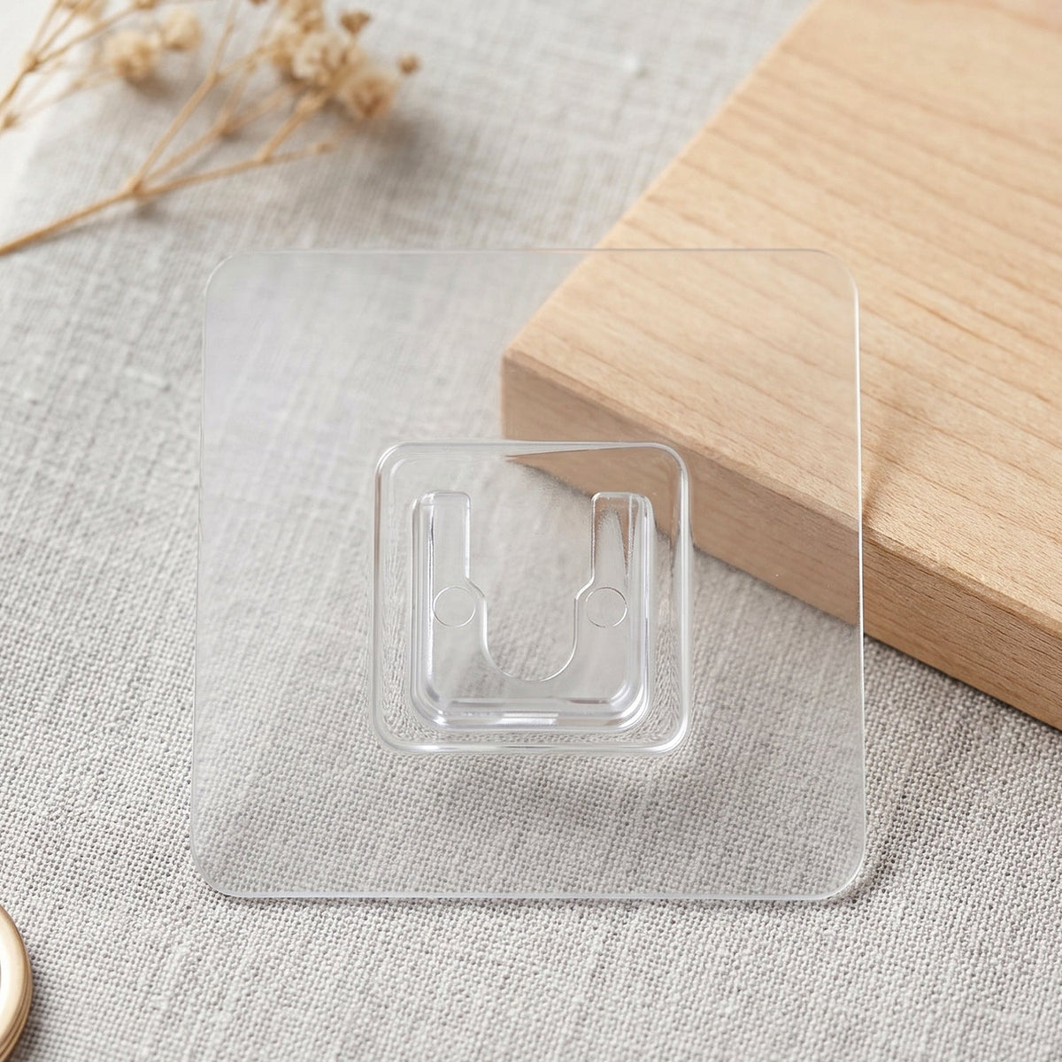 A transparent wall hook is displayed on a light gray textured surface with a wooden block and dried flowers in the