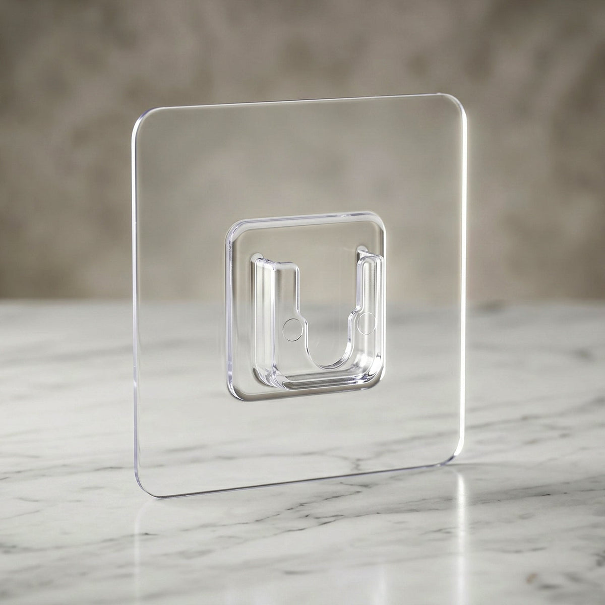 A clear, square self-adhesive hook stands upright on a white marble surface against a blurred gray background.