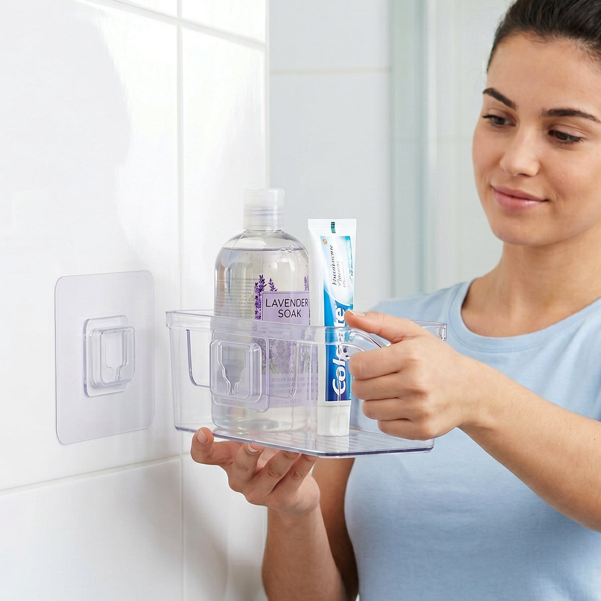 A woman places a clear caddy with shampoo and toothpaste onto a clear wall hook on a white tiled bathroom wall.