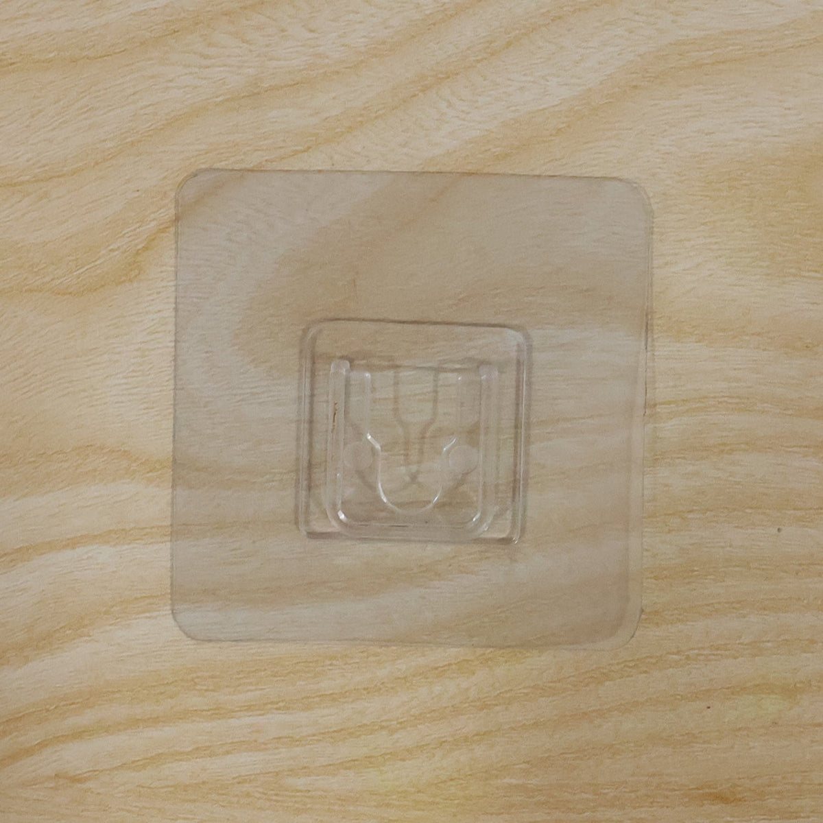 A clear, square self-adhesive hook is centered on a light brown wooden surface with a visible grain.