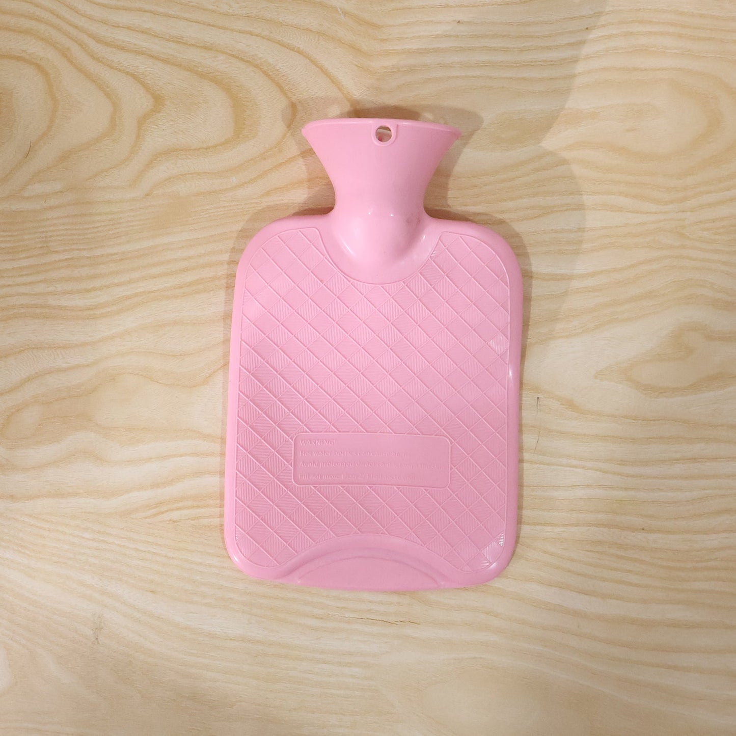 A pink hot water bag with a textured, diagonal stripe pattern, lying flat on a light wooden surface.