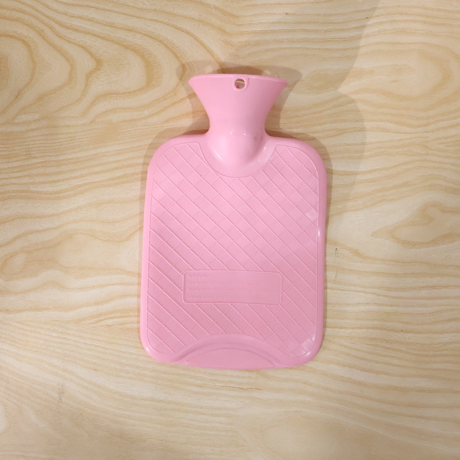 A pink hot water bag with a textured, diagonal stripe pattern, lying flat on a light wooden surface.