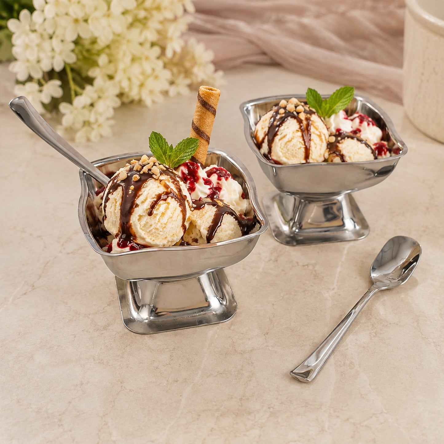 Two stainless steel ice cream cups filled with vanilla ice cream, chocolate sauce, and mint leaves, with a spoon.