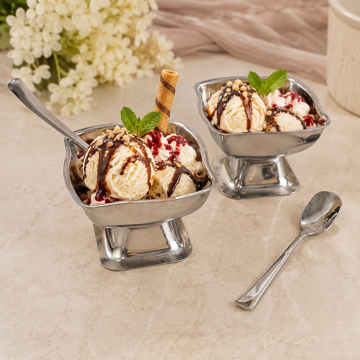 Two stainless steel ice cream cups filled with vanilla ice cream, chocolate sauce, and mint leaves, with a spoon.