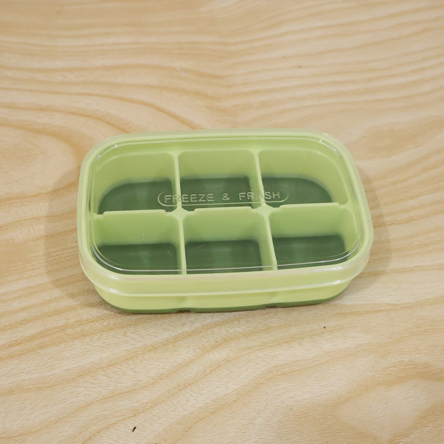 A light green 6-cavity ice cube tray with a clear lid, featuring dark green silicone bottoms, resting on a light wooden