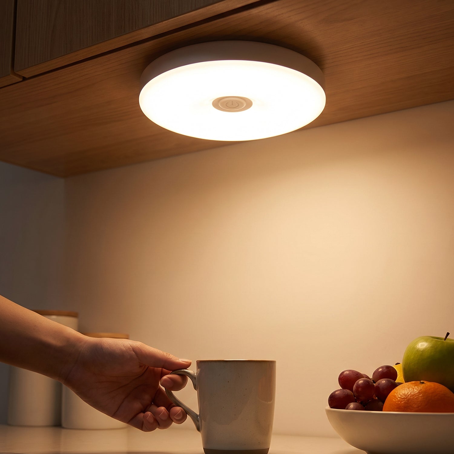 A hand reaches for a mug under a bright, round LED light mounted beneath a wooden cabinet, illuminating a fruit bowl