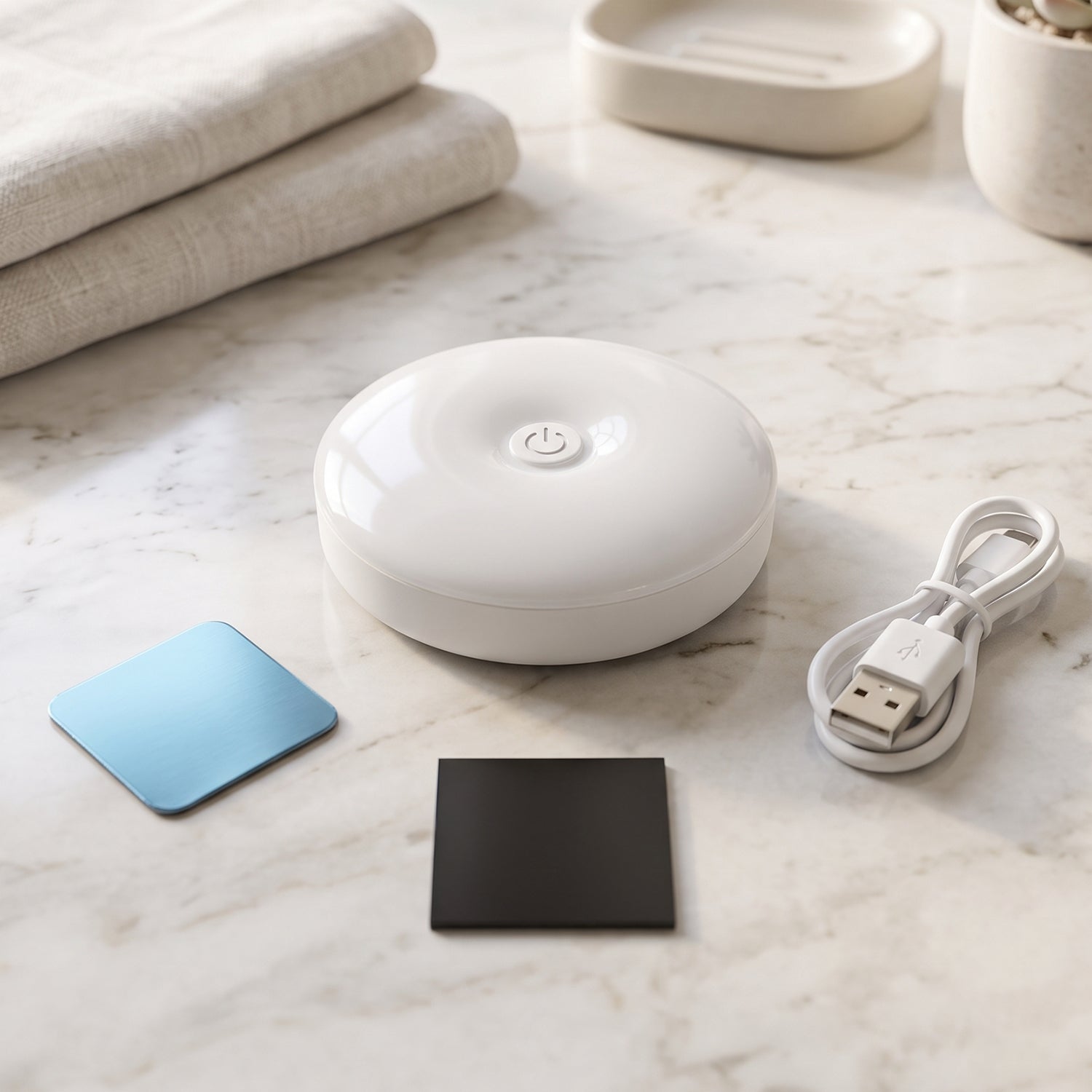 A white, round LED light, a white USB charging cable, a blue adhesive pad, and a black magnetic pad on a marble surface