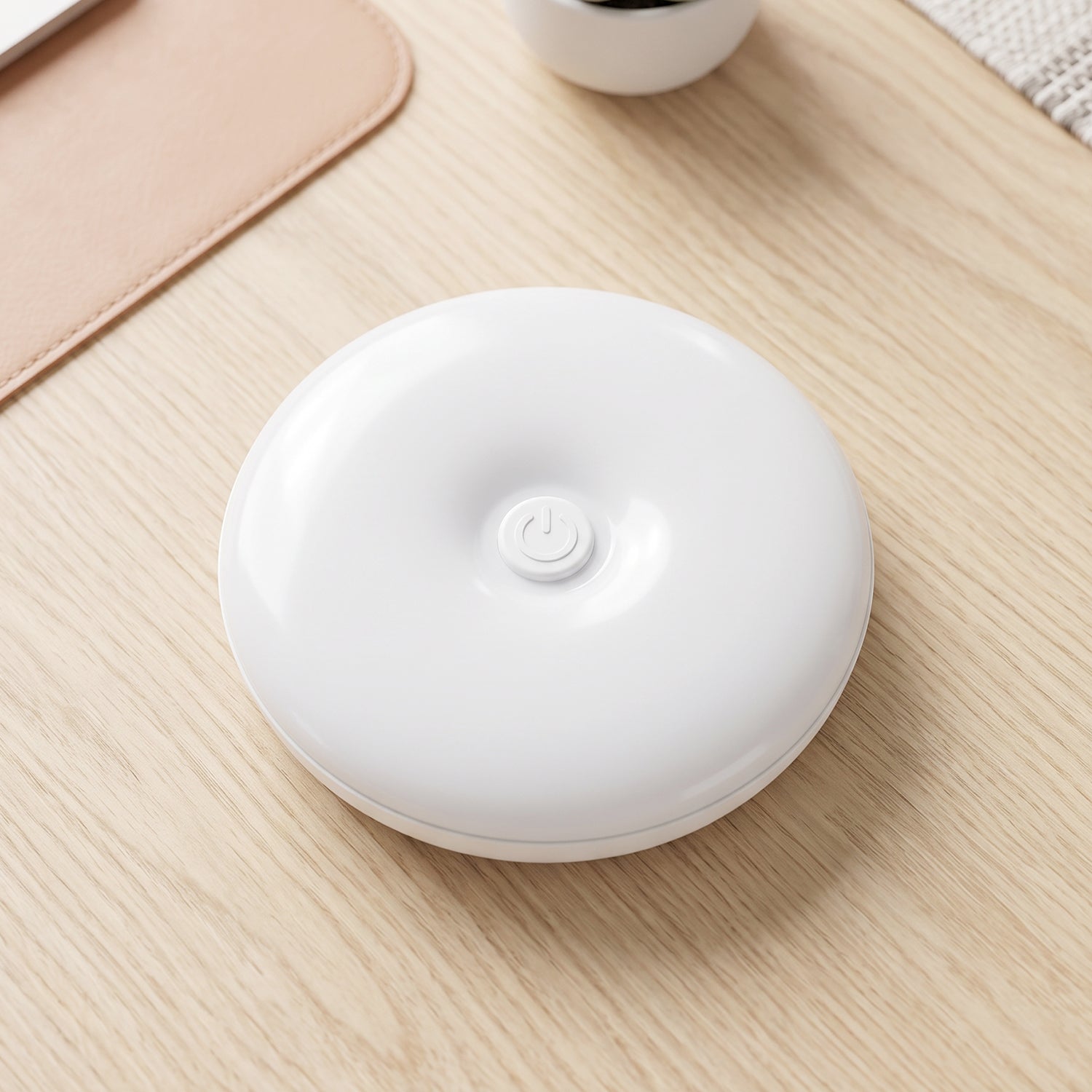 A white, round LED light with a central power button rests on a light wood desk next to a pink mat