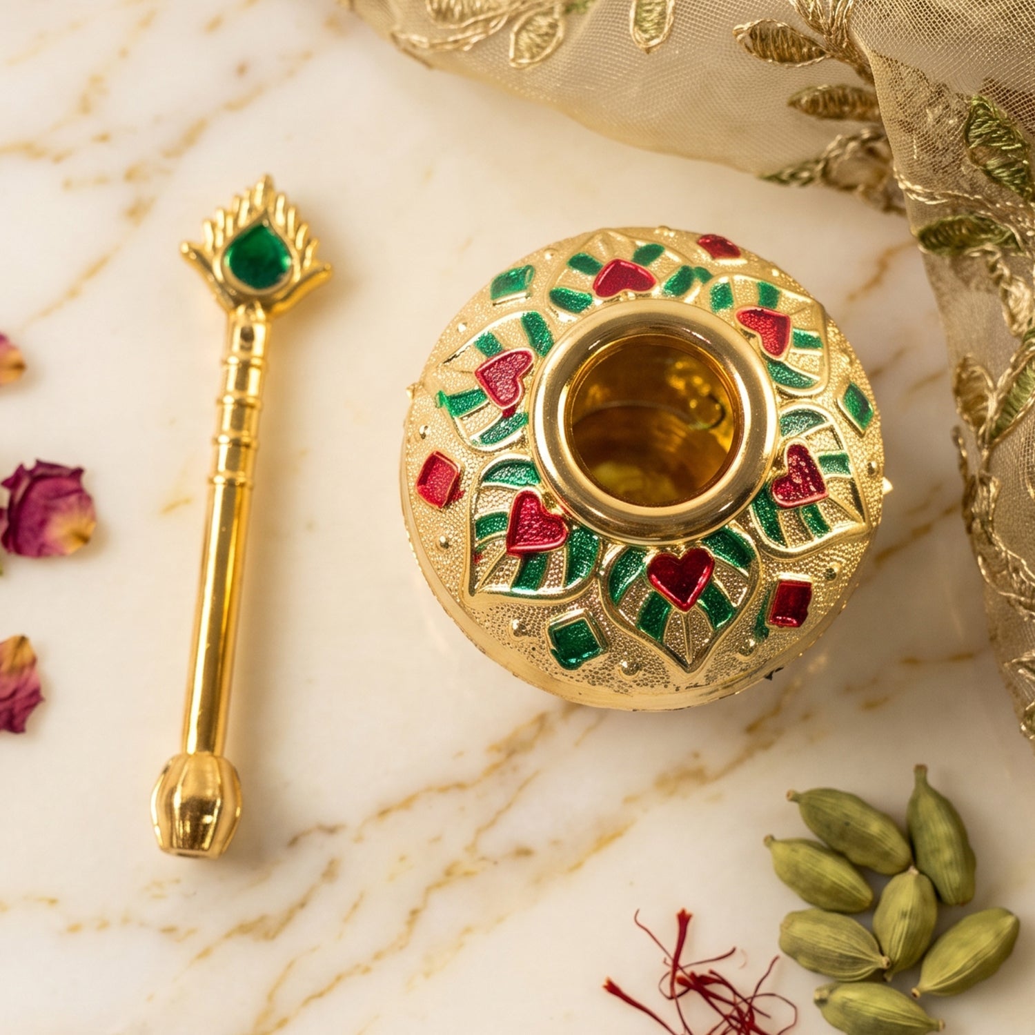 The gold decorative pot and spoon, both with red and green enamel, are displayed on a white marble surface with spices.