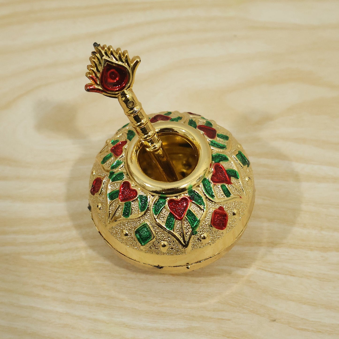 A gold decorative pot and spoon, with red and green enamel, are shown on a light wooden surface.