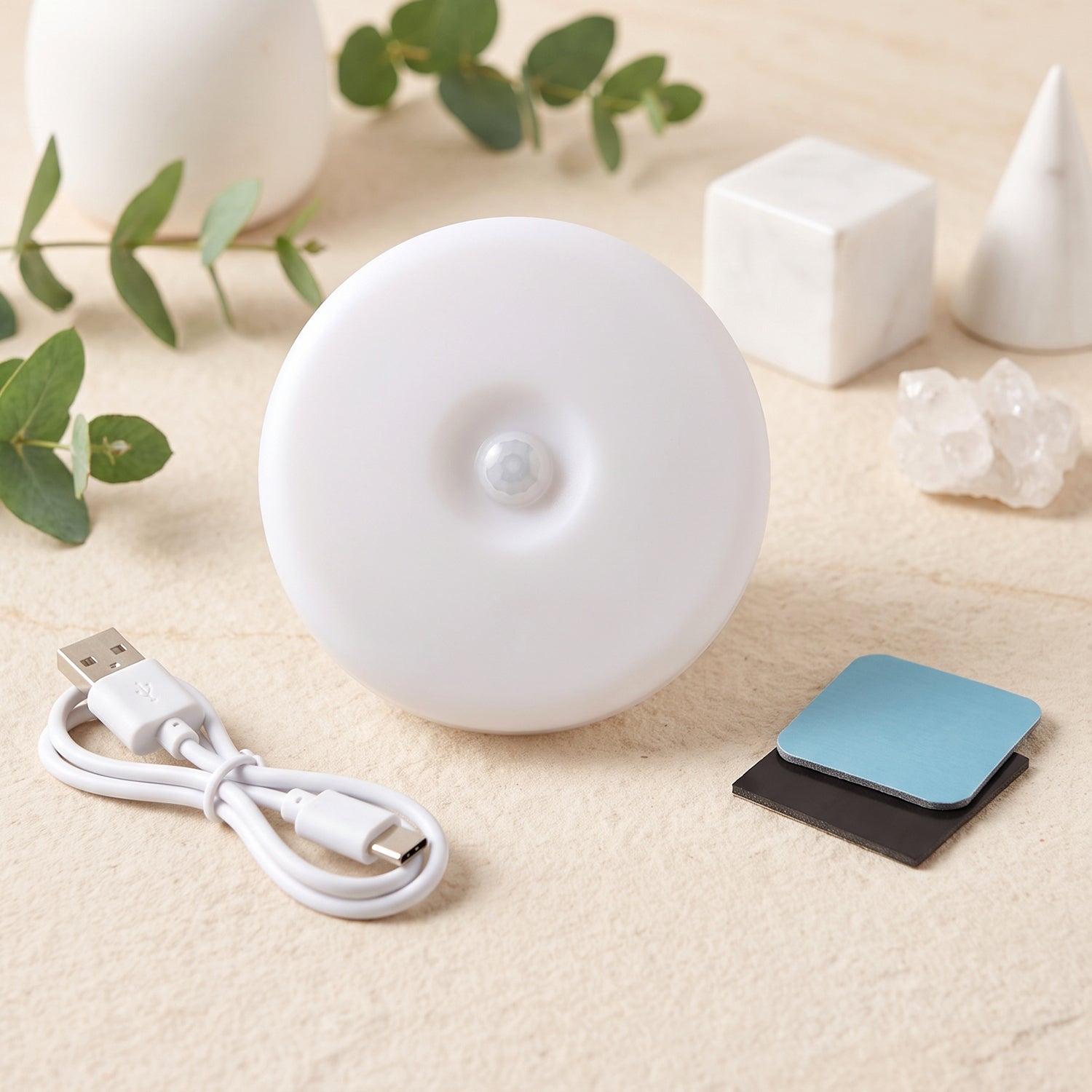 The white circular LED light with a central motion sensor is displayed with its USB charging cable and adhesive pads.
