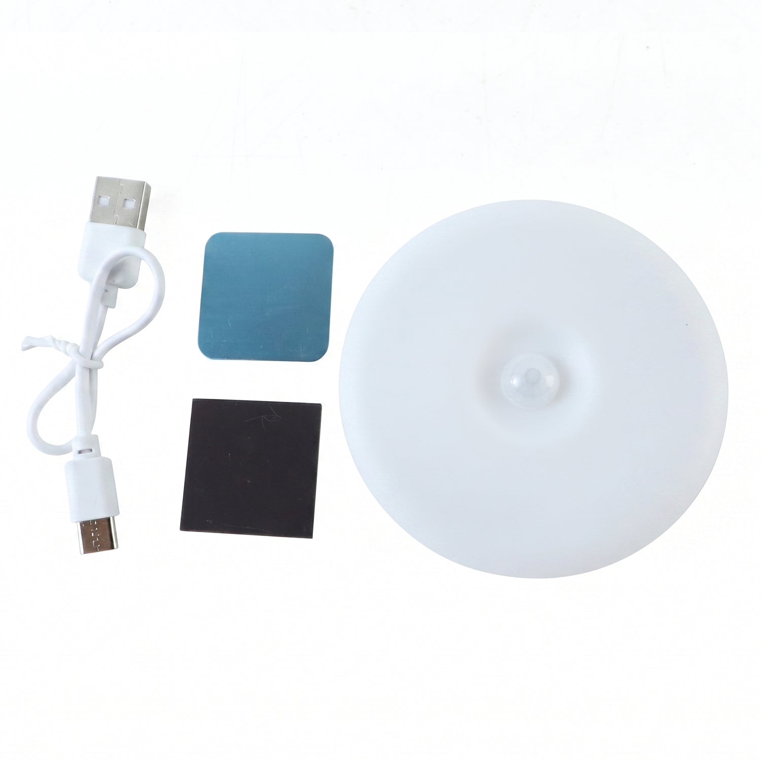 The white circular LED light, USB charging cable, and two adhesive pads are arranged on a plain white surface.