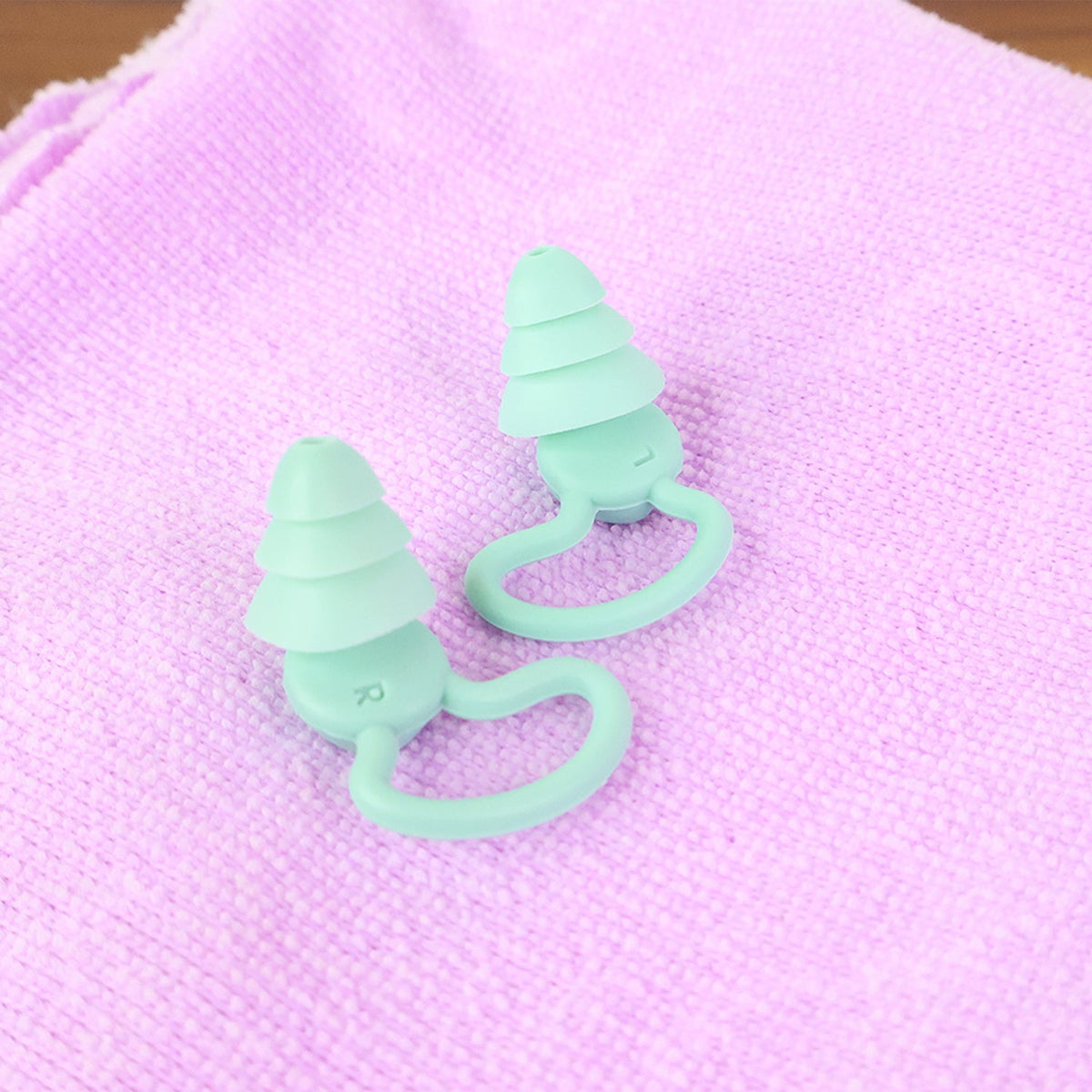 Two mint green silicone earplugs with ear hooks are placed on a textured light purple towel.
