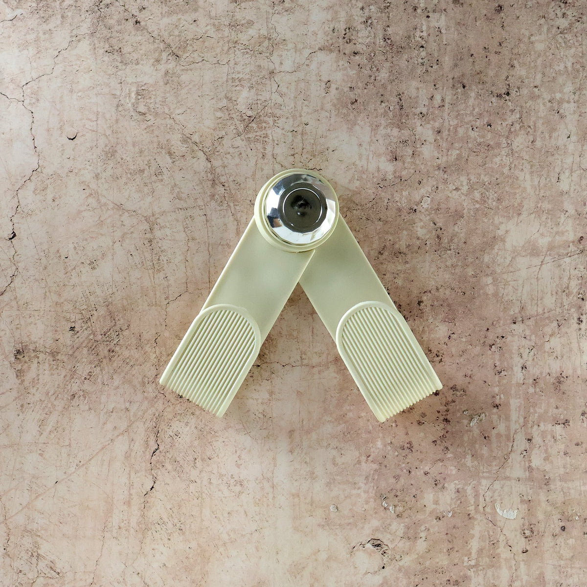A cream-colored, V-shaped pot lid holder with a chrome center is displayed against a textured, light brown background.