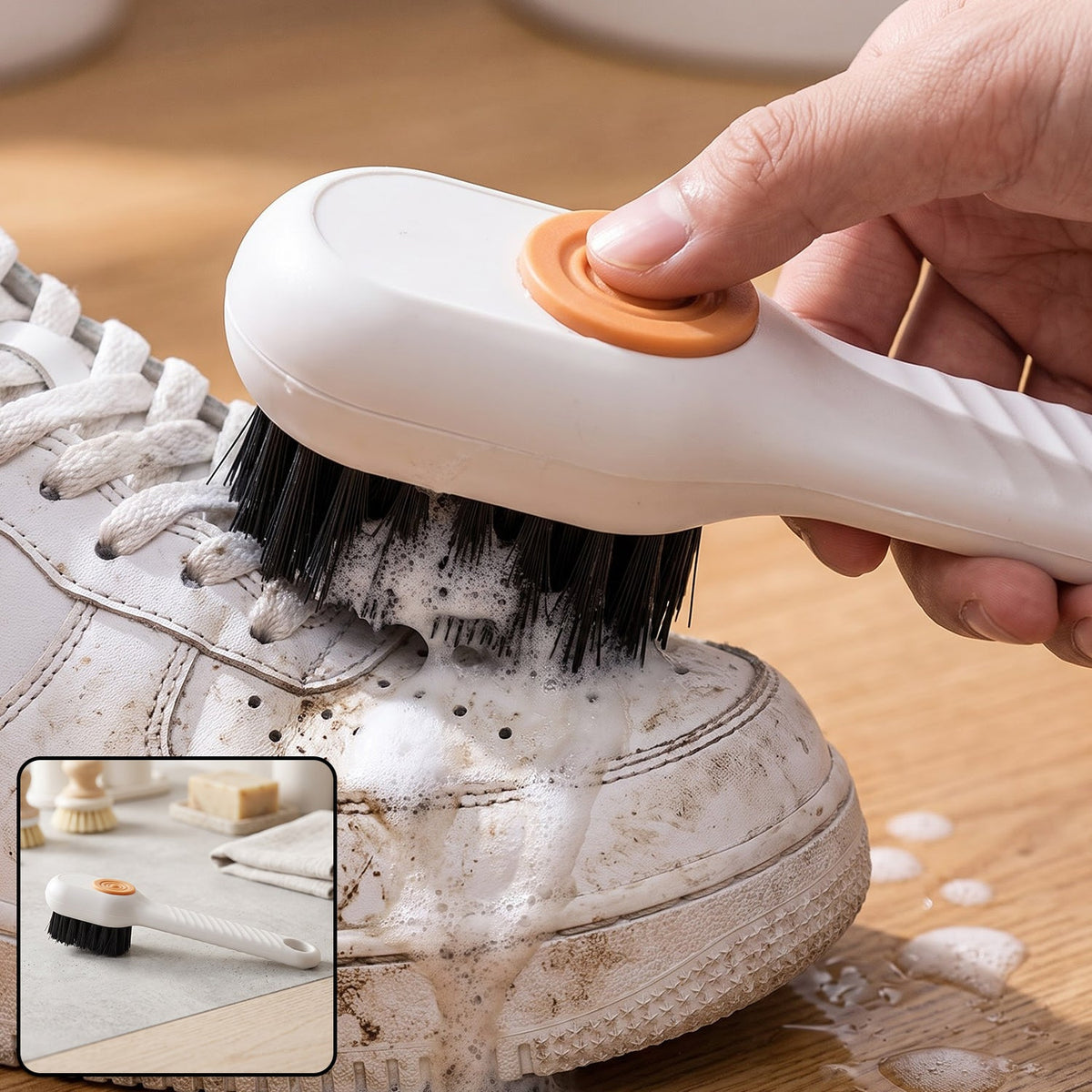 A hand holds a white scrubbing brush with an orange button, cleaning a dirty white sneaker with suds.
