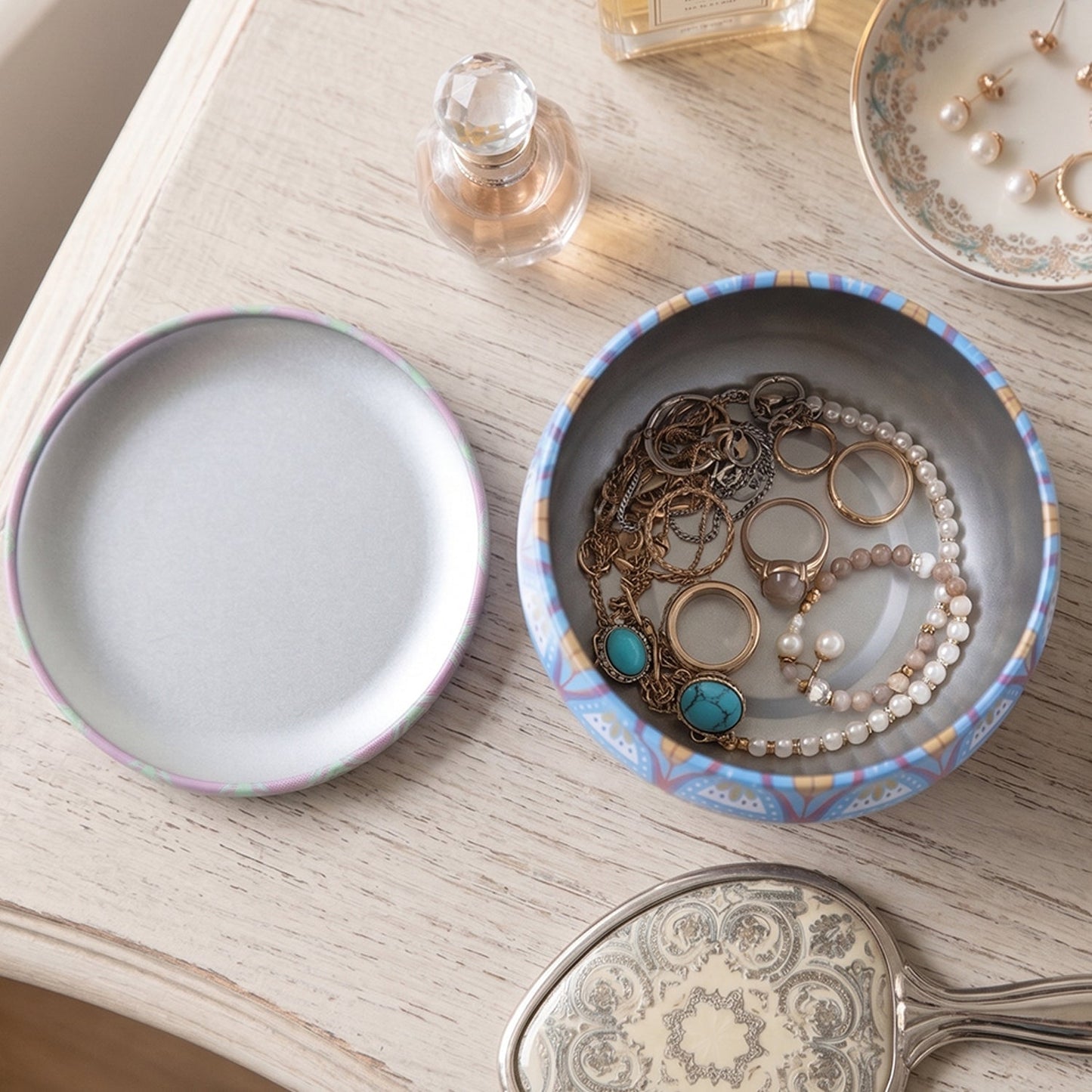 A mandala tin box filled with various jewelry pieces sits open next to its lid on a light wooden surface.