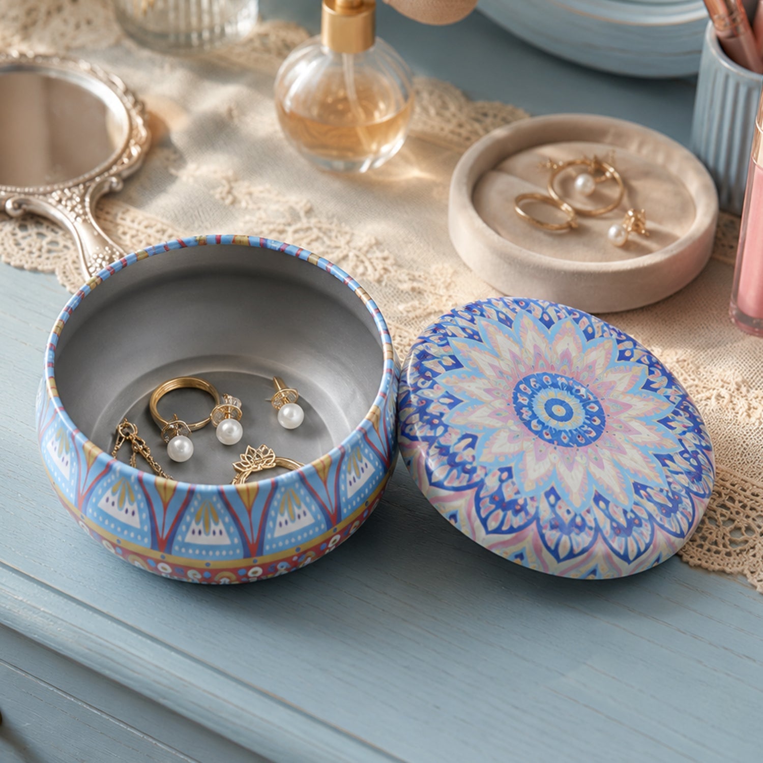 A decorative blue and orange mandala tin box, open with jewelry inside, and its lid resting on a blue lace cloth.