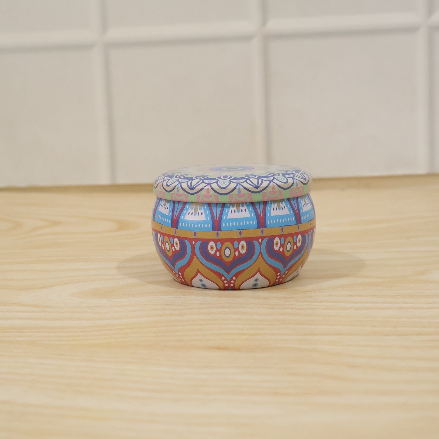 A small, closed blue and orange mandala tin box sits on a light wooden surface in front of a white tiled wall.