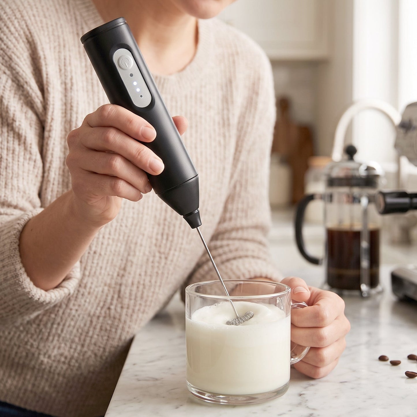 A person in a cream sweater uses a black electric milk frother to create foam in a clear glass of milk on a kitchen counter.