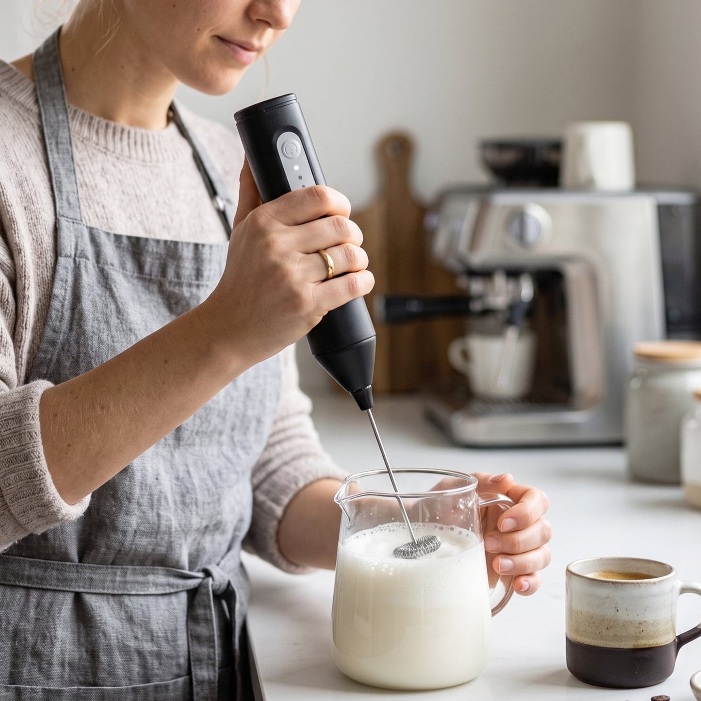 A person in a gray apron froths milk in a glass pitcher with a black electric frother, next to a two-toned coffee mug.