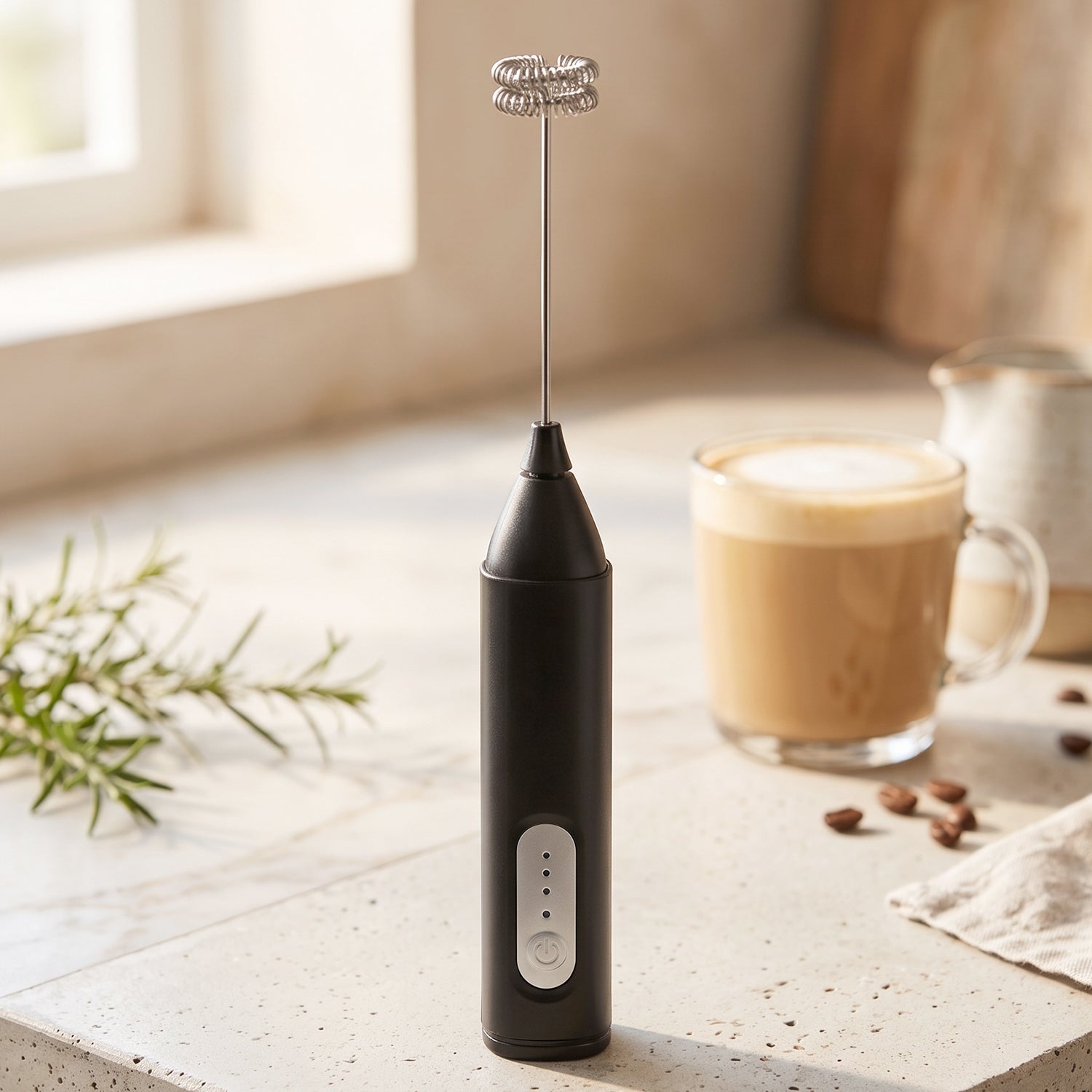 A black handheld electric milk frother stands on a light countertop with a latte in a glass mug and coffee beans in the