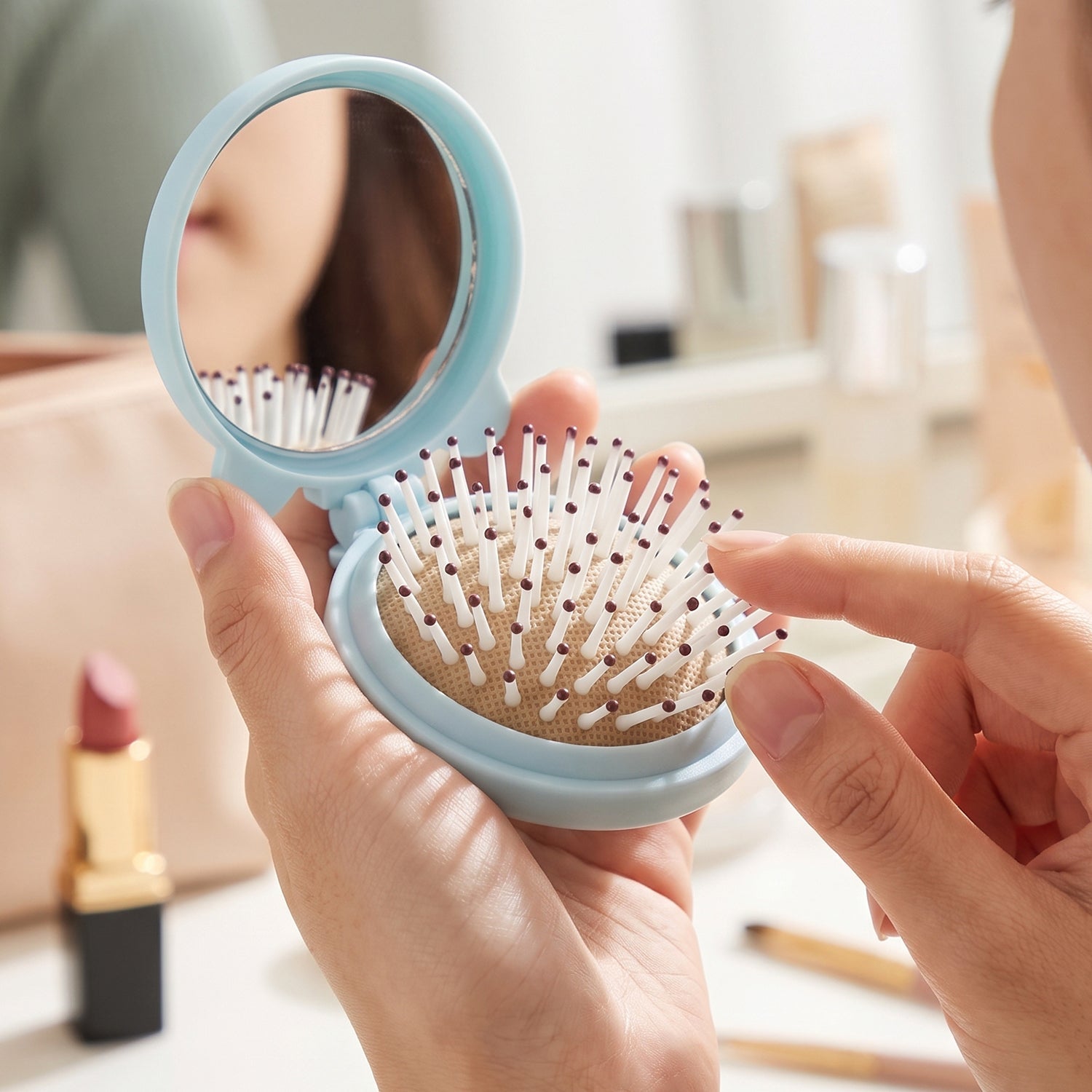 A person's hands hold a light blue folding hairbrush with a mirror, their finger touching the white and brown bristles.