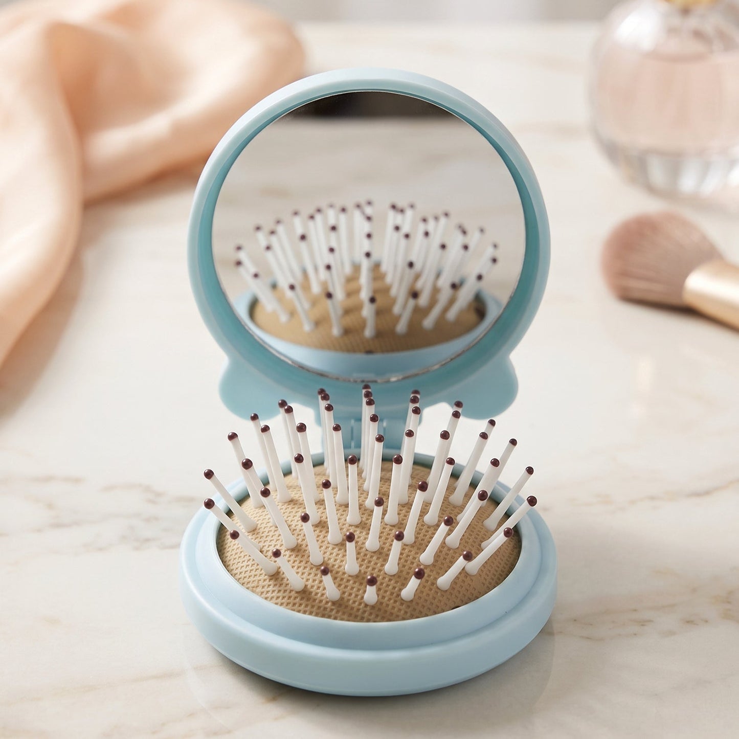 A light blue folding hairbrush with a mirror stands open on a marble surface, with a perfume bottle and brush in the soft