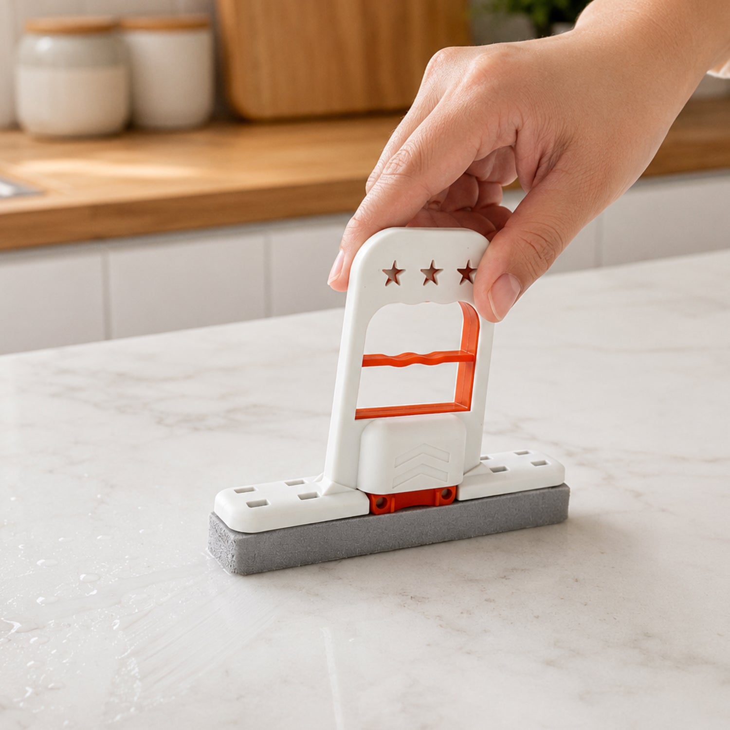 A hand holds a white and orange mini mop with a gray sponge, cleaning a white marble kitchen countertop.