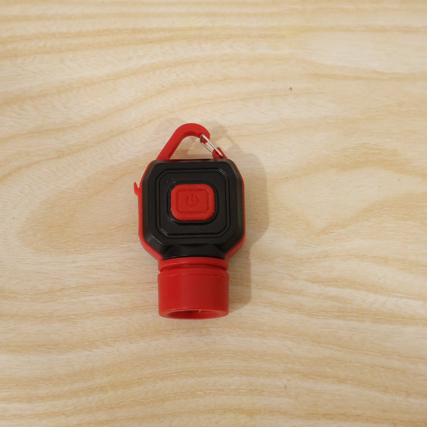 The back of the red and black mini LED flashlight with a red power button, lying on a light wooden surface.