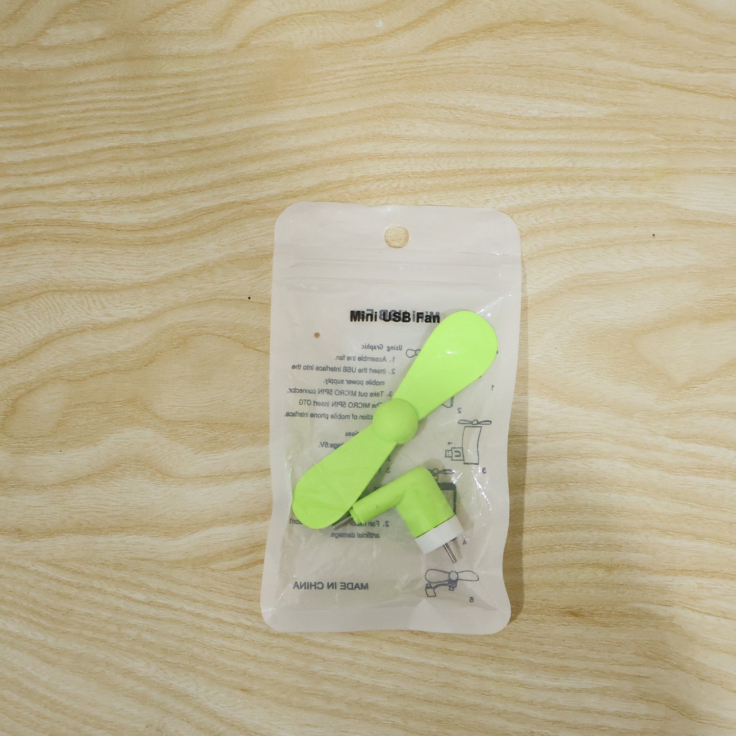 A bright green USB-C fan is sealed in clear plastic packaging on a light wooden surface.