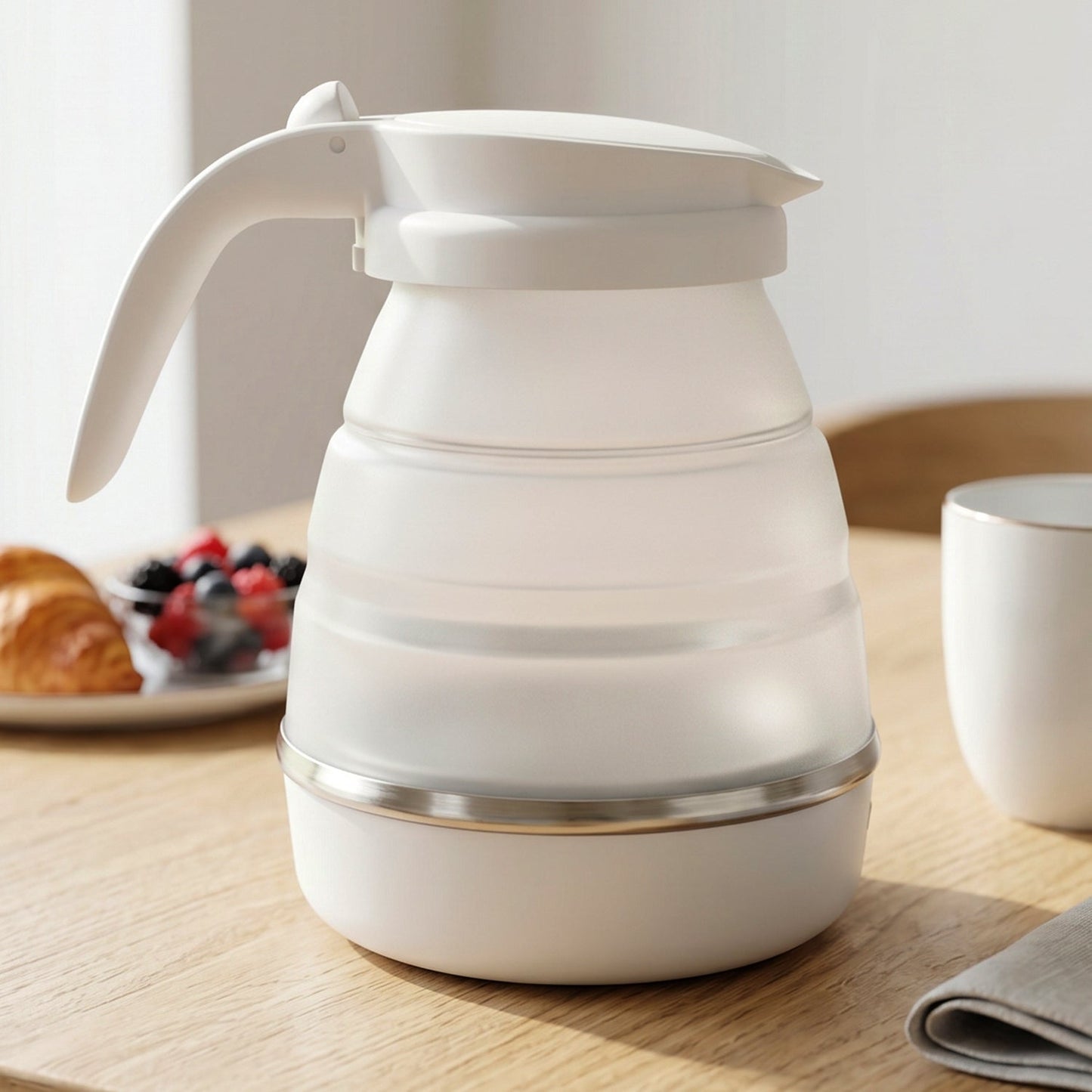 A white collapsible electric kettle on a wooden table, with a croissant and berries in a bowl in the background.