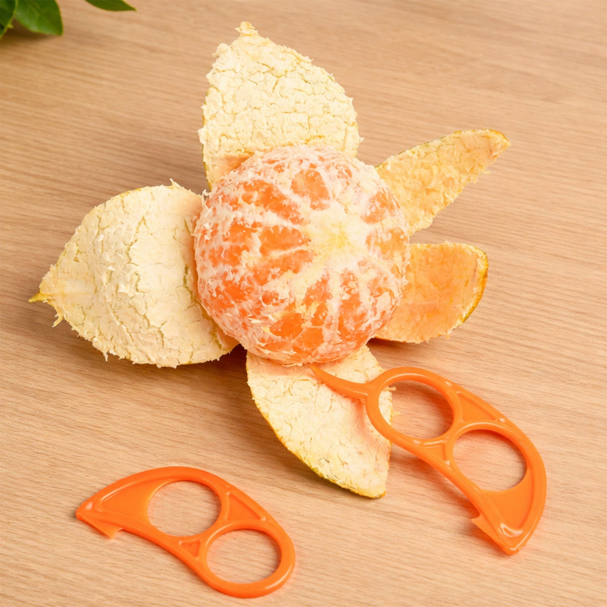 A partially peeled orange sits on a wooden table next to two orange plastic citrus peelers.