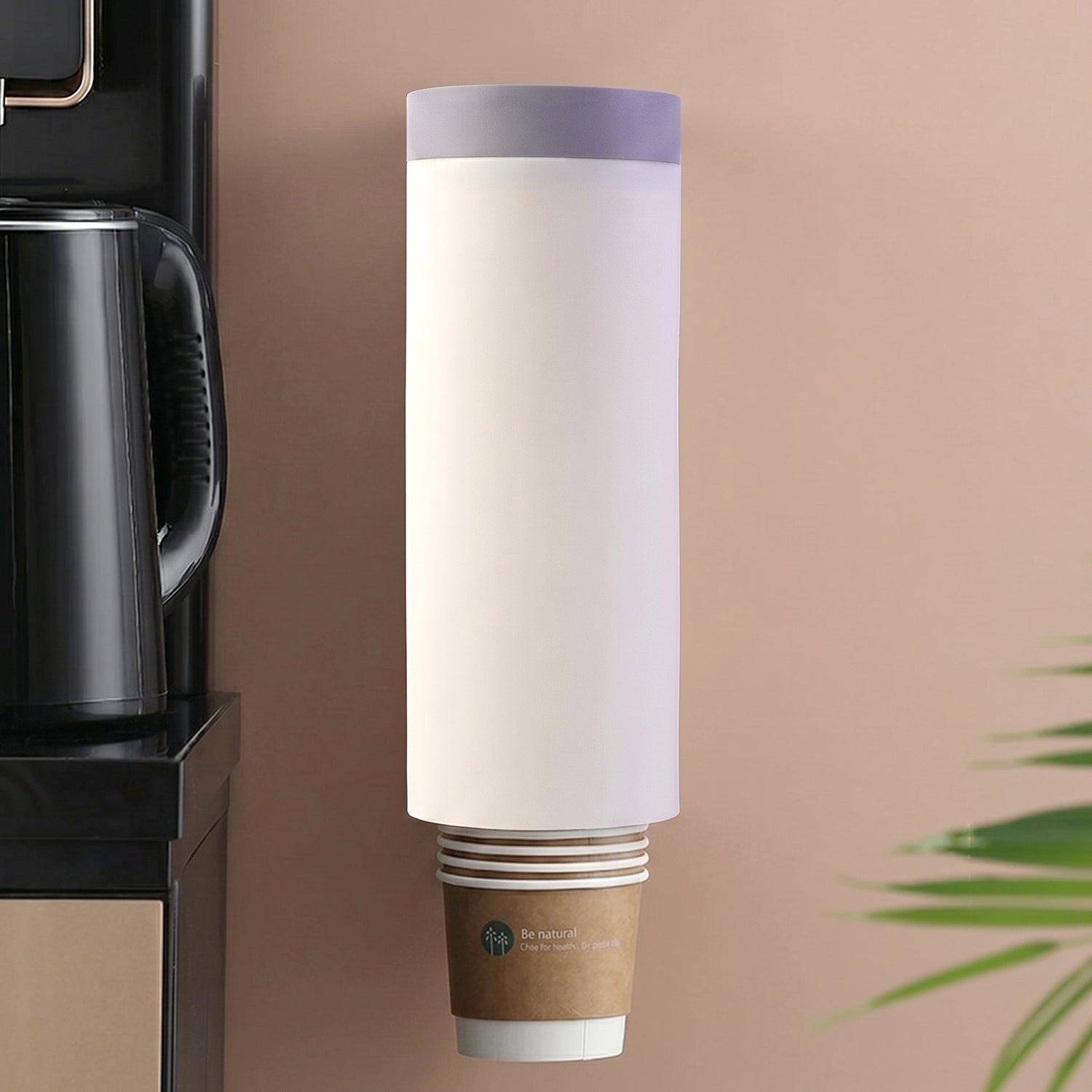 A white wall-mounted cup dispenser with a gray lid, holding a stack of brown paper cups, next to a black kettle.