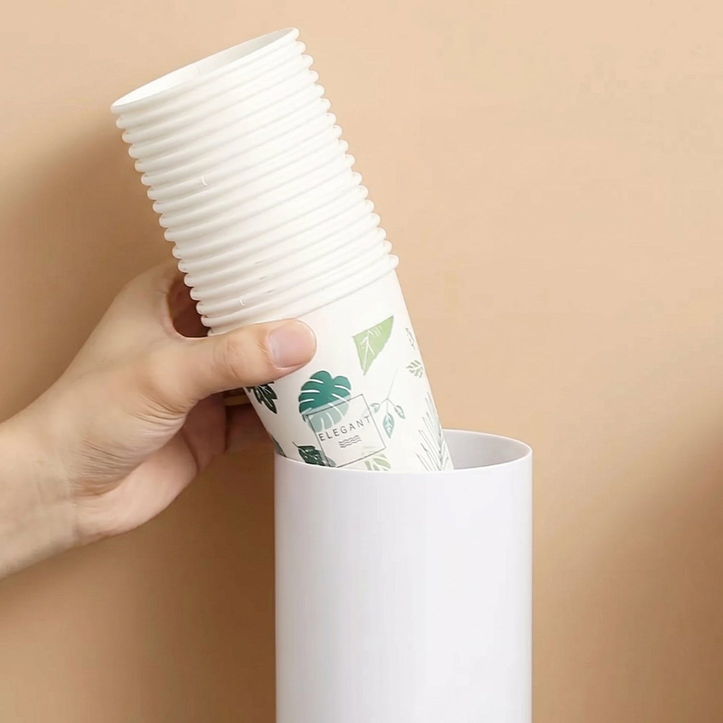 A hand inserting a stack of white paper cups with green leaf patterns into the top of a white cup dispenser.