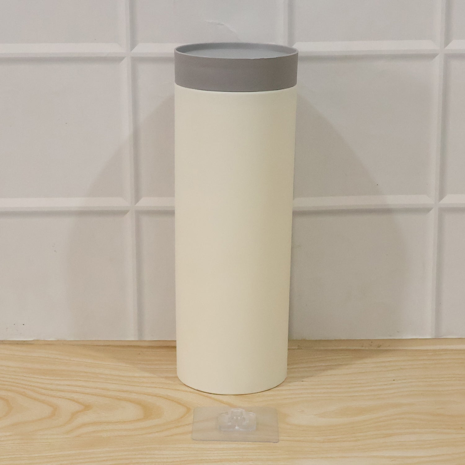 A white cylindrical cup dispenser with a gray lid, standing upright on a wooden surface in front of a white tiled wall, with