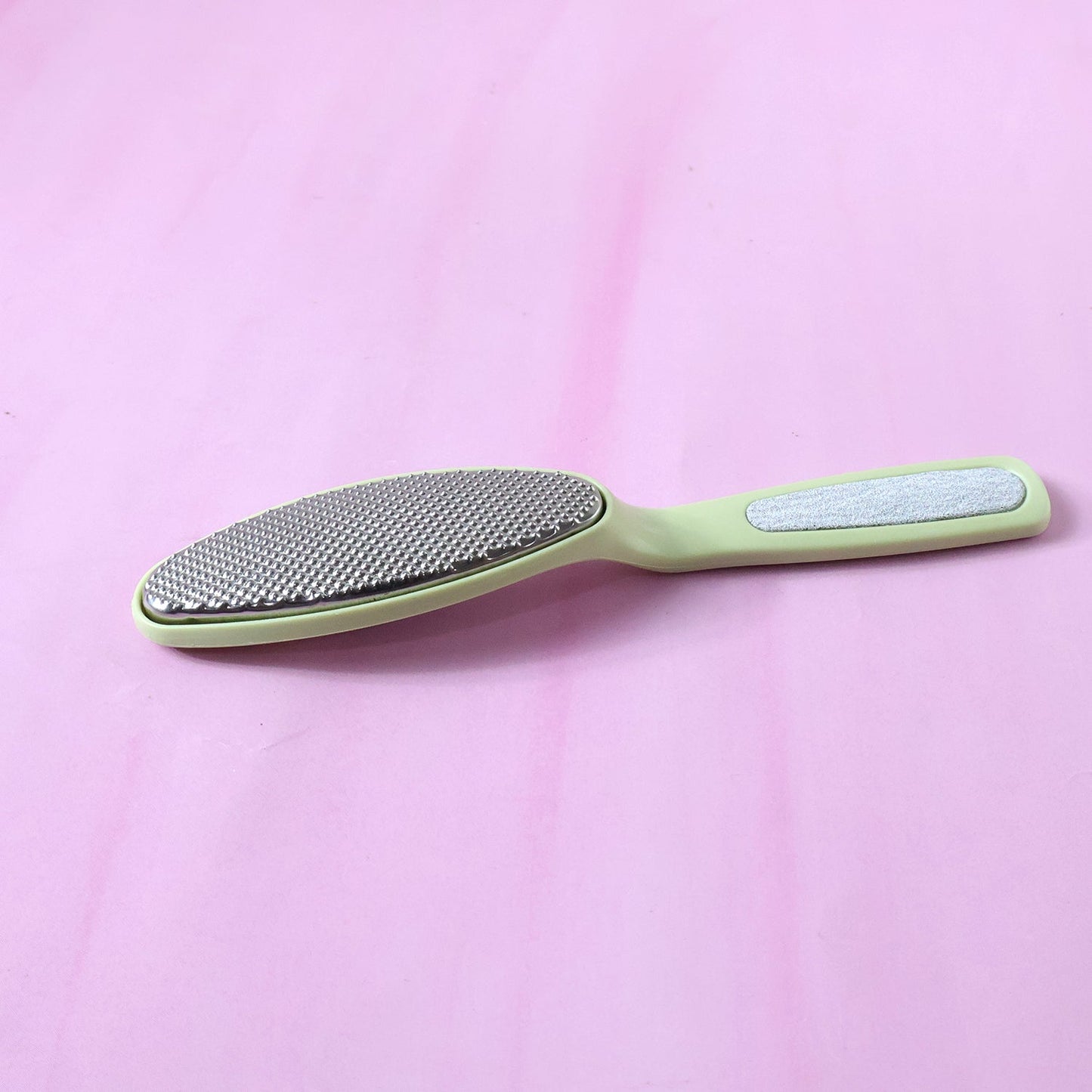 A light green dual-sided foot file with a silver textured surface and a smooth silver strip, against a pink background.