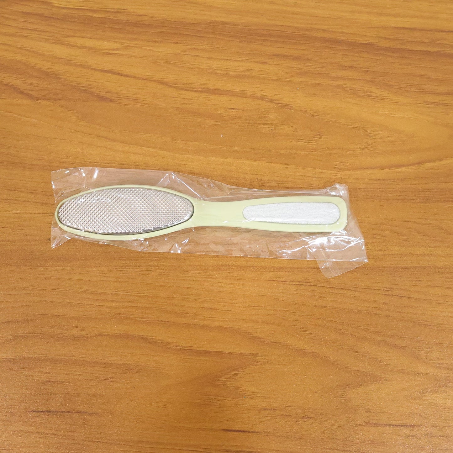 A light green dual-sided foot file, still sealed in clear plastic packaging, rests on a wooden surface.