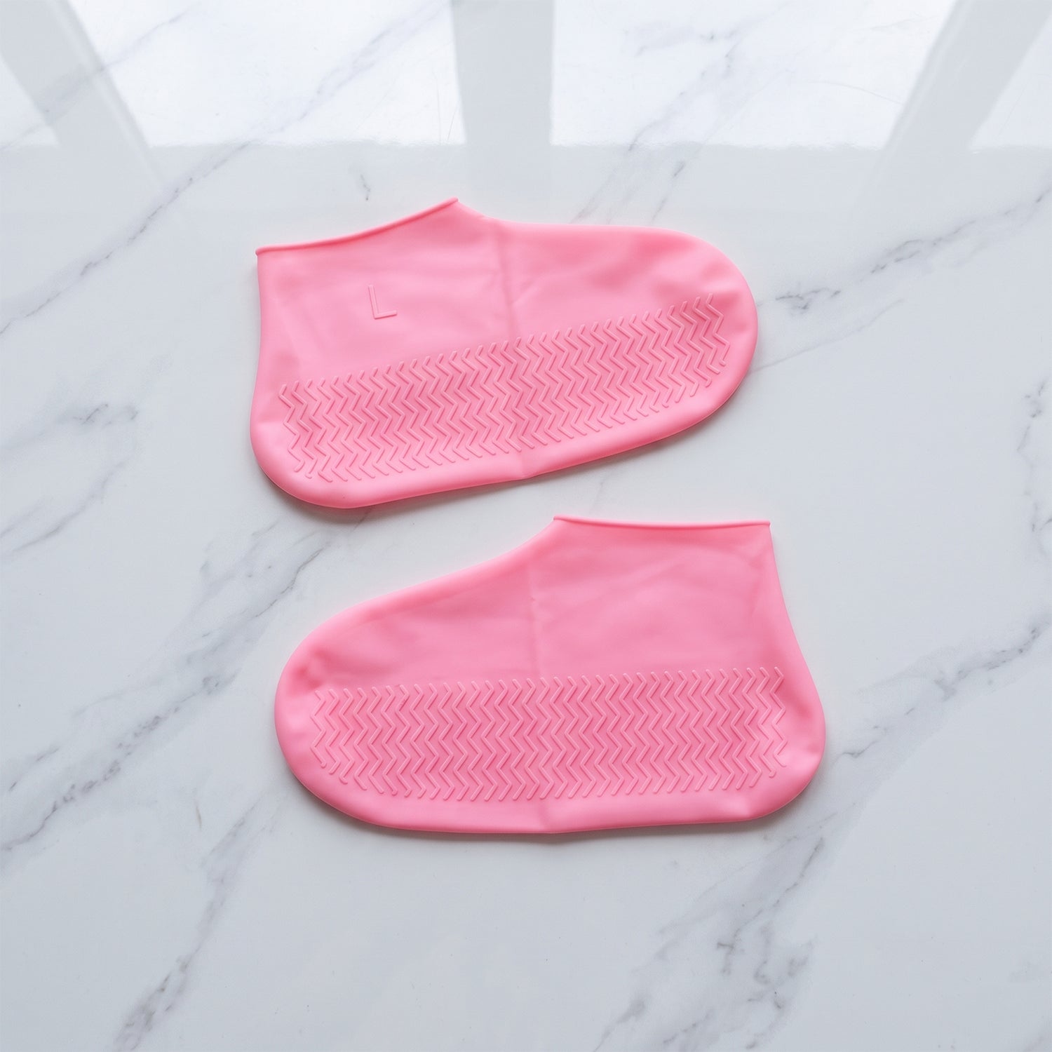 A pair of pink silicone shoe covers, marked with an L, lies flat on a white marble surface, displaying their textured soles.