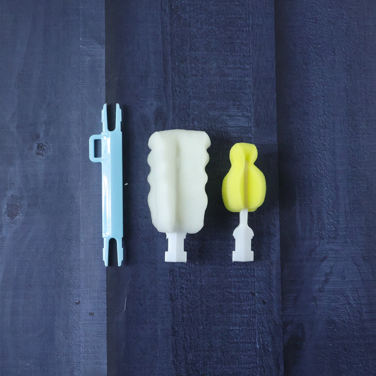 The disassembled parts of the cleaning brush: a light blue handle, a white sponge head, and a yellow sponge head, on a dark